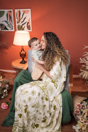 A woman with curly hair embraces a young child while sitting on a chair in a cozy, warmly lit room with a rustic, natural decor theme. The woman is wearing a long, patterned dress in green and white. Beside them is a table with a lit lamp, and floral arrangements decorate the surroundings.