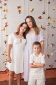 A woman and two children stand in front of a floral-decorated wall. The woman and an older girl with long hair, both in light-colored dresses, stand closely together, while a young boy in a white outfit stands with his arms crossed. The backdrop features hanging flowers of various colors such as red, white, peach, and green.