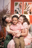 A joyful scene with three people closely embracing each other on a couch. The person on the left is wearing a yellow-striped shirt, the person in the center is in a pink polo shirt, and the person on the right is dressed in a pink dress. They are all smiling and radiating happiness. In the background, there are artistic wall decorations with abstract designs and a soft lamp providing warm lighting.