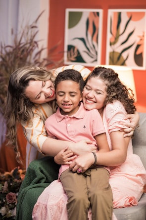 A joyful scene with three people closely embracing each other on a couch. The person on the left is wearing a yellow-striped shirt, the person in the center is in a pink polo shirt, and the person on the right is dressed in a pink dress. They are all smiling and radiating happiness. In the background, there are artistic wall decorations with abstract designs and a soft lamp providing warm lighting.