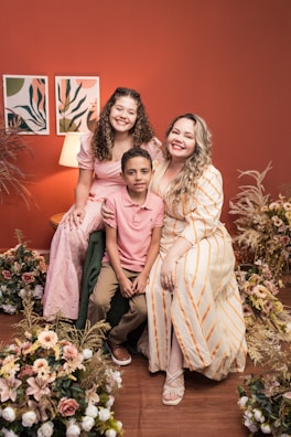 A joyful family portrait featuring three people sitting together. They are surrounded by floral arrangements. The background includes a warm red wall with two art pieces and a lit lamp, creating a cozy atmosphere.