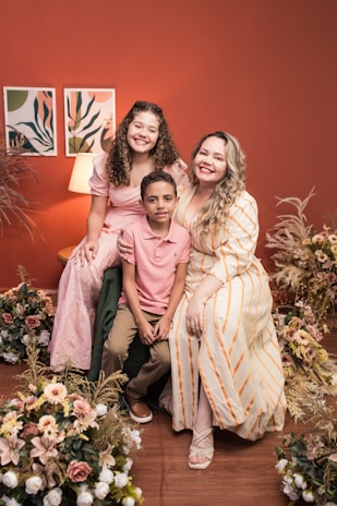 A joyful family portrait featuring three people sitting together. They are surrounded by floral arrangements. The background includes a warm red wall with two art pieces and a lit lamp, creating a cozy atmosphere.