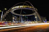 A large illuminated architectural structure with the words 'biswa bangla gate' is prominently displayed. The structure is ring-shaped and elevated over a roadway, surrounded by light trails from passing vehicles. The scene is set at night, with nearby buildings and streetlights visible.