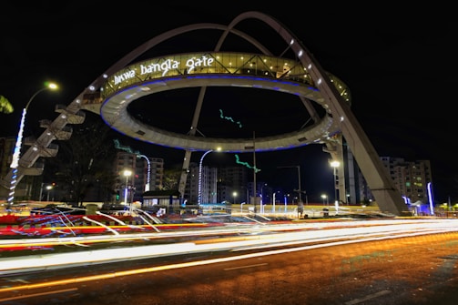 A large illuminated architectural structure with the words 'biswa bangla gate' is prominently displayed. The structure is ring-shaped and elevated over a roadway, surrounded by light trails from passing vehicles. The scene is set at night, with nearby buildings and streetlights visible.