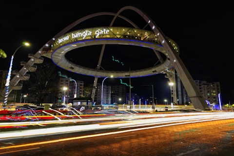 A large illuminated architectural structure with the words 'biswa bangla gate' is prominently displayed. The structure is ring-shaped and elevated over a roadway, surrounded by light trails from passing vehicles. The scene is set at night, with nearby buildings and streetlights visible.