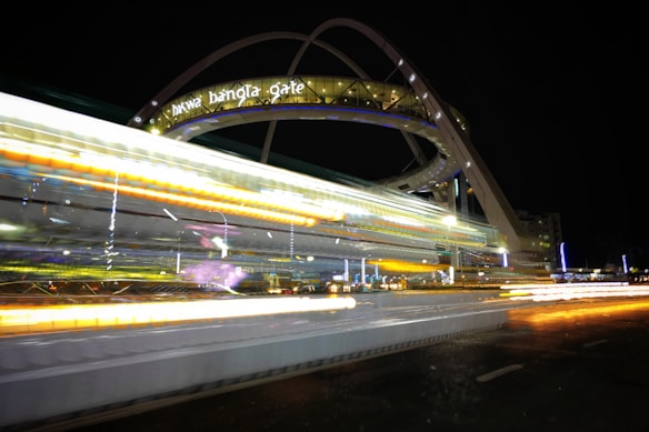 An architectural structure with an illuminated sign reading 'HITWA BANGLA GATE' is captured at night. The image features bright and colorful light trails, suggesting long-exposure photography, indicating fast-moving vehicles in the foreground. The structure appears to be a modern gateway or arch with dynamic curves.