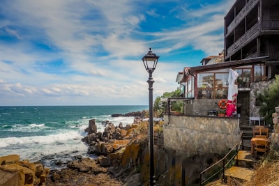 A coastal scene featuring a house built on a rocky shoreline. The building has stone walls, large windows, and a balcony overlooking the ocean. A streetlamp stands nearby, and a lifebuoy is visible on the wall. Waves crash against the rocks under a partly cloudy sky.