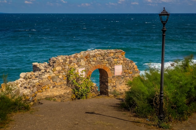 A stone archway stands on a cliff overlooking the deep blue ocean. The rustic arch is surrounded by green shrubs and a weathered lamp post. The sky is dotted with light clouds, and the sunlight illuminates the stone, highlighting its earthy tones.