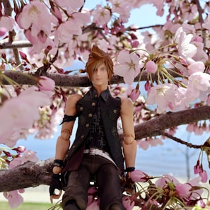 A detailed anime-style figurine is seated on a tree branch surrounded by blossoming pink cherry blossoms. The figurine wears a dark vest and pants, featuring intricate designs on its outfit. The background includes a clear blue sky and a hint of a distant, blurred landscape.