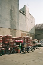 A stack of beverage crates, primarily red and white, is organized against a large, weathered wall in an open area. A small vendor stall with a bright pink cover is partially visible among the crates. Two bicycles and three-wheel carts are parked nearby. In the background, two ambulances are parked near another building with a partially visible damaged roof.