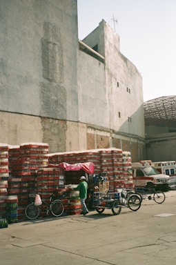 A stack of beverage crates, primarily red and white, is organized against a large, weathered wall in an open area. A small vendor stall with a bright pink cover is partially visible among the crates. Two bicycles and three-wheel carts are parked nearby. In the background, two ambulances are parked near another building with a partially visible damaged roof.