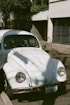 A vintage white Volkswagen Beetle is parked on a street. The car has visible signs of wear and rust on the bumper. Shadows of nearby trees fall across the car's surface. There is a building with a metal gate in the background, and the scene is illuminated by natural daylight.