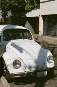 A vintage white Volkswagen Beetle is parked on a street. The car has visible signs of wear and rust on the bumper. Shadows of nearby trees fall across the car's surface. There is a building with a metal gate in the background, and the scene is illuminated by natural daylight.