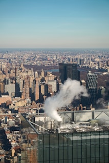 A densely packed urban skyline with numerous high-rise buildings, reflecting sunlight on their glass surfaces. A plume of white steam rises prominently from the rooftop of one of the buildings in the foreground, against a clear blue sky.
