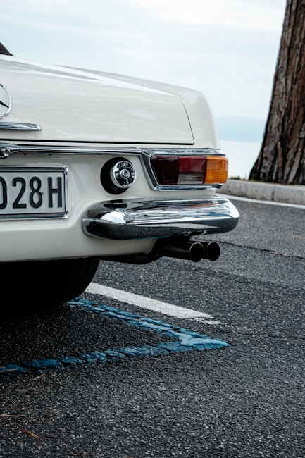 A vintage white car is parked on a textured asphalt surface, featuring a detailed rear view with chrome elements, a rectangular license plate, and protruding exhaust pipes. A tree trunk and a hazy horizon can be seen in the background.