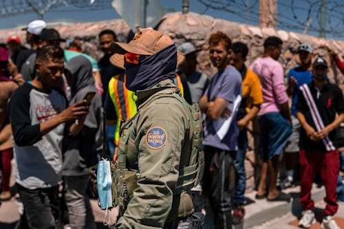A U.S. Border Patrol officer stands in the foreground, wearing a khaki uniform and protective gear, while a group of individuals stand behind a fence in the background. The scene includes people wearing casual clothing, some holding documents, and a barbed wire fence. The officer appears to be engaged in monitoring or communication.