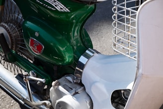 A close-up of a vintage-style motorcycle with a green body and distinctive logo. The image highlights the details of the engine casing, chrome handle, metal spokes, and part of a white front guard. The chrome exhaust pipe and wire headlight guard are also visible.