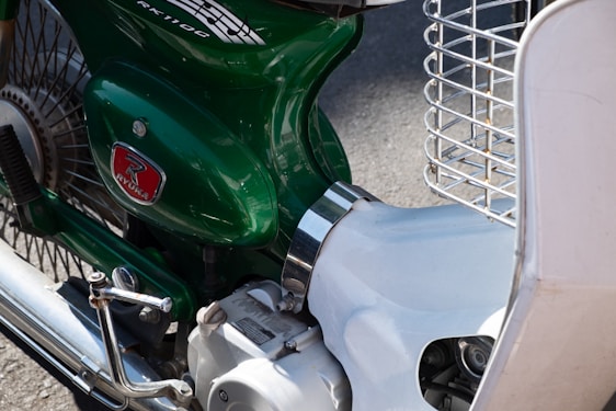 A close-up of a vintage-style motorcycle with a green body and distinctive logo. The image highlights the details of the engine casing, chrome handle, metal spokes, and part of a white front guard. The chrome exhaust pipe and wire headlight guard are also visible.
