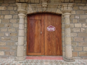 A wooden double door is framed by stone columns and a stone wall, featuring an arched design above. A sign on the door displays operating hours from 7:00 am to 12:00 noon and 2:00 pm to 7:00 pm. The floor in front of the door has a patterned tile design.