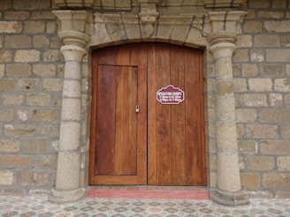 A wooden double door is framed by stone columns and a stone wall, featuring an arched design above. A sign on the door displays operating hours from 7:00 am to 12:00 noon and 2:00 pm to 7:00 pm. The floor in front of the door has a patterned tile design.