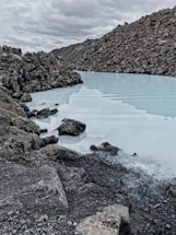 A serene landscape featuring a milky blue geothermal pool surrounded by rugged volcanic rocks. The cloudy sky adds to the dramatic and tranquil atmosphere. The stark contrast between the smooth water surface and the rough texture of the rocks creates a visually striking scene.