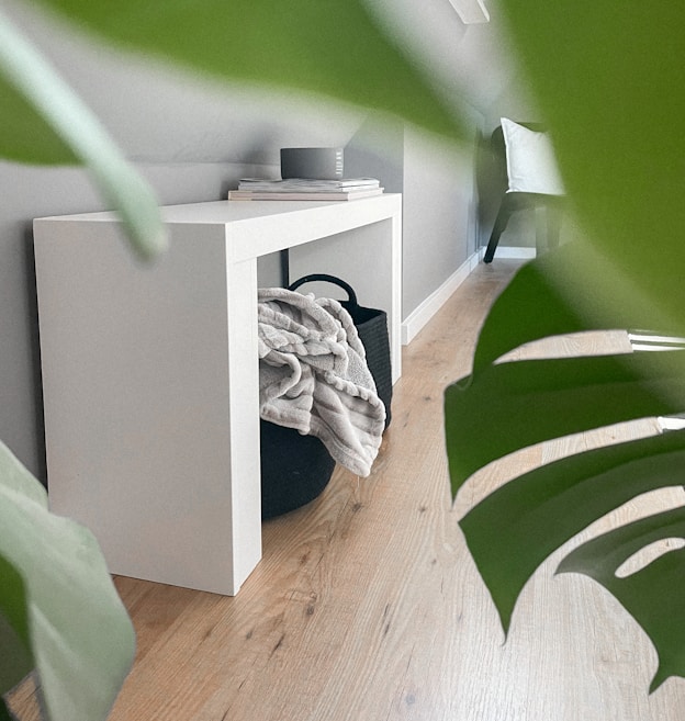 A minimalist interior scene featuring a sleek, white console table situated against a gray wall. On top of the table are a small stack of books and a cylindrical object. Beneath the table is a black basket containing a neatly folded blanket. The floor is wooden, adding warmth to the setting, and large green leaves partially frame the image, suggesting the presence of indoor plants. In the background, there is a chair with a white cushion.