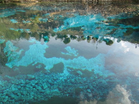 A serene body of water with clear reflections of surrounding trees and clouds. The water is crystal clear, showing vibrant blue and turquoise tones with visible stones at the bottom. The scenery reflects a peaceful and natural landscape.