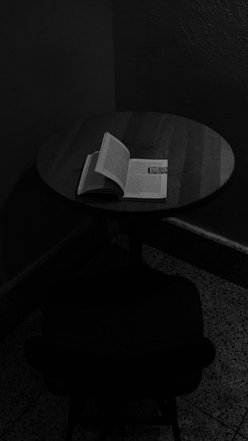 A dimly lit scene features a round wooden table with an open book resting on it. Shadows cast across the floor and wall create a moody atmosphere.