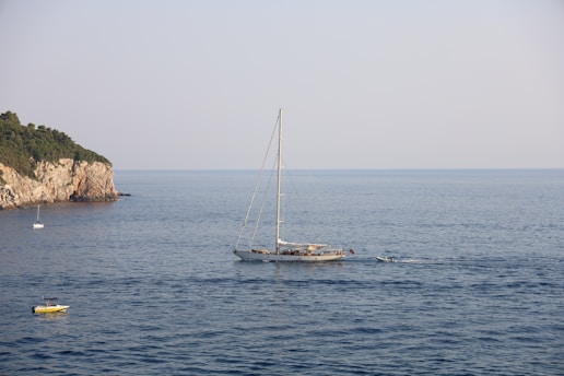 A large sailboat is in the center of the image, gliding across calm blue waters near a rocky coastline. The sky is clear with a light haze. A small island or cliff covered with greenery is visible to the left, and two smaller boats can be seen in the foreground and background.