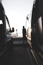 A cinematic golden hour shot of a man standing beside his van parked on a rugged mountain trail.