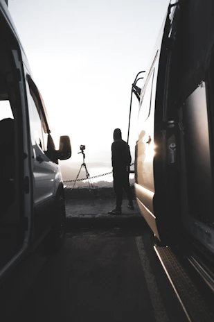A cinematic golden hour shot of a man standing beside his van parked on a rugged mountain trail.