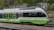 A modern passenger train with a sleek, aerodynamic design is stationed on the railway tracks. It is predominantly green and gray with the logo 'transN' visibly displayed on its side. The background consists of lush greenery and a rock wall.