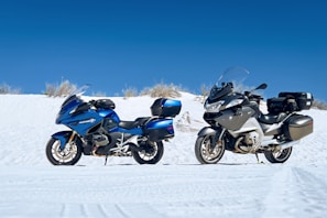 Two motorcycles are parked side by side on a white sandy terrain under a clear blue sky. The motorcycle on the left is metallic blue with black accents, while the one on the right is silver and black. Both vehicles are equipped with windshields and storage cases.