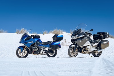 Two motorcycles are parked side by side on a white sandy terrain under a clear blue sky. The motorcycle on the left is metallic blue with black accents, while the one on the right is silver and black. Both vehicles are equipped with windshields and storage cases.