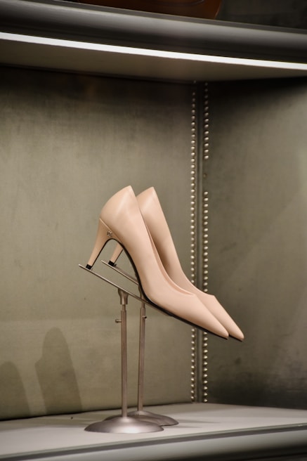 A pair of elegant high-heeled shoes displayed on a metal stand against a soft, green textured background with metallic studs outlining the edge.