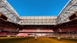 An expansive indoor soccer stadium with an open roof, revealing a bright blue sky. The seating areas are primarily red, and the field surface appears green with artificial lighting structures positioned above. The stadium features modern architecture with large screens and intricate metal framing.