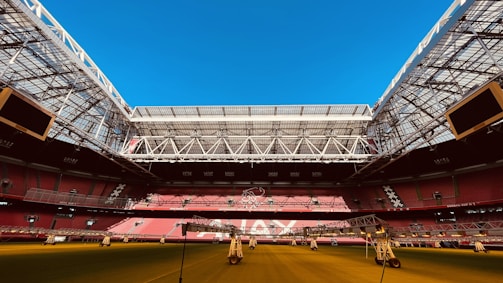 An expansive indoor soccer stadium with an open roof, revealing a bright blue sky. The seating areas are primarily red, and the field surface appears green with artificial lighting structures positioned above. The stadium features modern architecture with large screens and intricate metal framing.