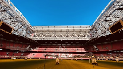 An expansive indoor soccer stadium with an open roof, revealing a bright blue sky. The seating areas are primarily red, and the field surface appears green with artificial lighting structures positioned above. The stadium features modern architecture with large screens and intricate metal framing.