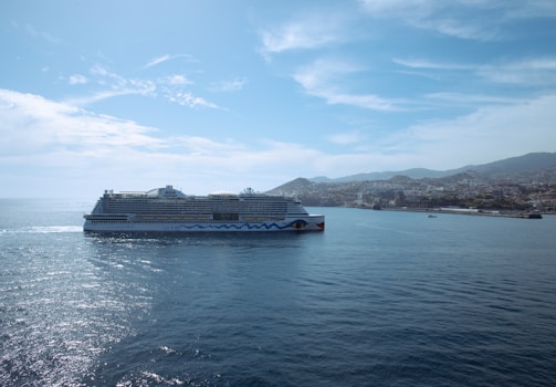 A large cruise ship sails on a calm sea near a coastal city with a mountainous backdrop. The sky is partly cloudy with patches of blue, casting a serene atmosphere over the scene.