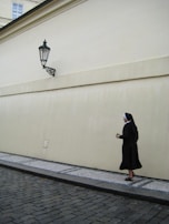 A nun dressed in a black habit and white coif walks along a cobblestone street next to a beige wall. The streetlamp attached to the wall casts a shadow over the pavement.