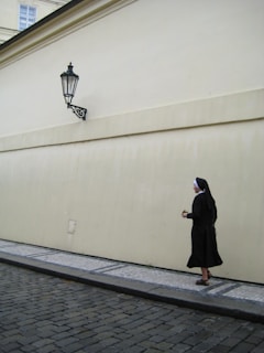 A nun dressed in a black habit and white coif walks along a cobblestone street next to a beige wall. The streetlamp attached to the wall casts a shadow over the pavement.