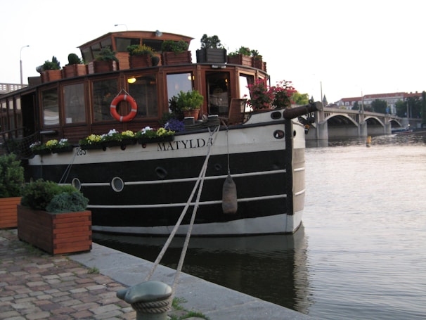 A charming vintage boat is docked by the riverside, with wooden siding and a small cabin adorned with numerous potted plants and flowers. A lifebuoy is attached near the windows, and there are ropes securing the boat to the dock. In the background, a bridge spans across the river, with buildings and faint cityscape visible beyond.