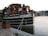 A charming vintage boat is docked by the riverside, with wooden siding and a small cabin adorned with numerous potted plants and flowers. A lifebuoy is attached near the windows, and there are ropes securing the boat to the dock. In the background, a bridge spans across the river, with buildings and faint cityscape visible beyond.