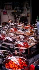 A market stall displaying a variety of colorful dried ingredients in metal bowls covered with transparent domes. The ingredients range in color from bright orange and deep red to earthy browns, indicating a selection of spices or preserved foods. The setting appears cluttered with numerous small containers, likely in an indoor or covered market environment.