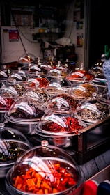 A market stall displaying a variety of colorful dried ingredients in metal bowls covered with transparent domes. The ingredients range in color from bright orange and deep red to earthy browns, indicating a selection of spices or preserved foods. The setting appears cluttered with numerous small containers, likely in an indoor or covered market environment.