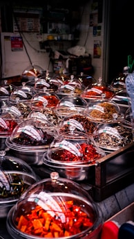 A market stall displaying a variety of colorful dried ingredients in metal bowls covered with transparent domes. The ingredients range in color from bright orange and deep red to earthy browns, indicating a selection of spices or preserved foods. The setting appears cluttered with numerous small containers, likely in an indoor or covered market environment.