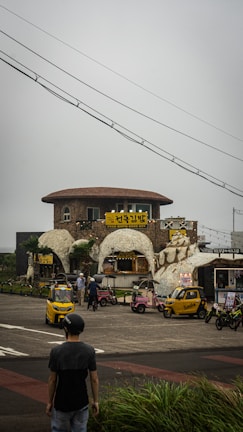A stone building with a rounded roof is surrounded by small, colorful vehicles parked outside. The signage is written in Korean. Several people are gathered around, with one person walking towards the area. Overhead, power lines stretch across the sky, and the overall scene has a cloudy backdrop.