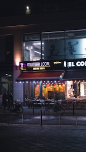 A storefront of a restaurant named 'Mumbai Local' offering Indian food is illuminated in the evening. Colorful lanterns hang from the ceiling, and the outdoor seating area is visible with tables and chairs arranged neatly. Bicycles are parked nearby, and a few pedestrians are seen walking past. The signage is vibrant, with a mix of English and possibly Hindi text.