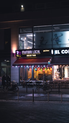 A storefront of a restaurant named 'Mumbai Local' offering Indian food is illuminated in the evening. Colorful lanterns hang from the ceiling, and the outdoor seating area is visible with tables and chairs arranged neatly. Bicycles are parked nearby, and a few pedestrians are seen walking past. The signage is vibrant, with a mix of English and possibly Hindi text.