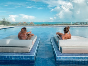 Two people are relaxing on lounge floats in a luxurious pool, with a scenic view of the ocean in the background. The sky is clear with a few clouds, and there are some thatched umbrellas visible in the distance. Both individuals are lounging back-to-back while enjoying drinks.